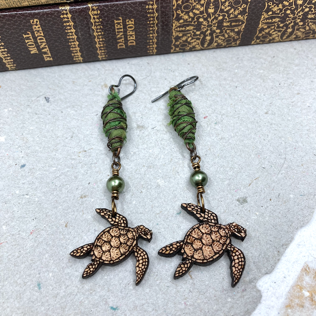 Earrings with patterned wooden turtle charms, green pearls and green fiber and wire cocoon connectors. 
