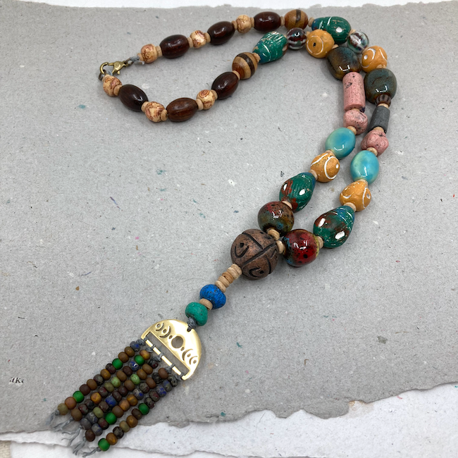 Necklace with gold charm with seed beads hanging off. Beads are a variety of shapes, colors, textures, and materials. 
